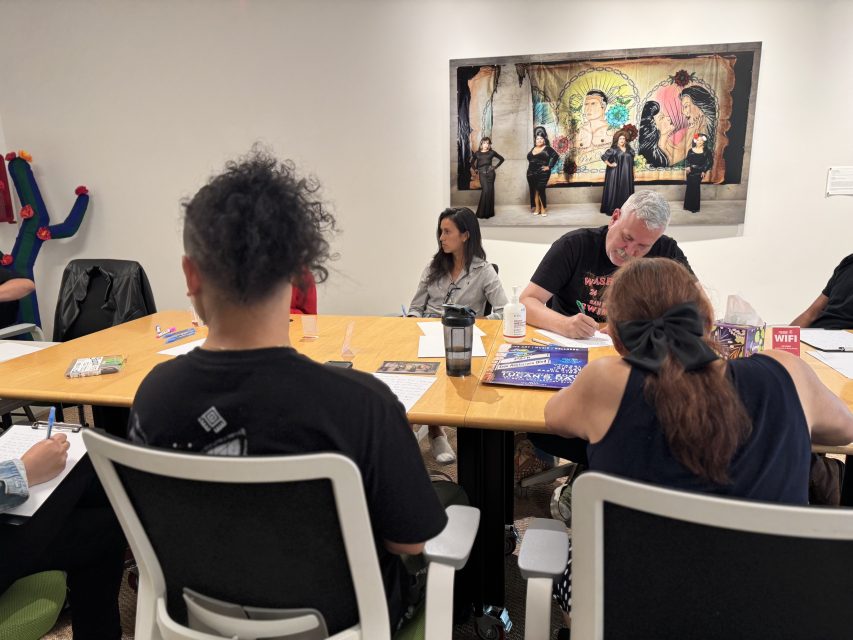 A group of people sit around a table engaged in a meeting or workshop; artwork with several figures is displayed on the wall behind them.