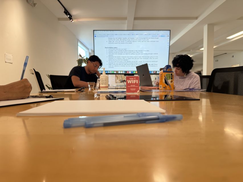 Three people work at a conference table with laptops and papers; a large screen displays text, and a red WiFi information card sits in the center of the table.