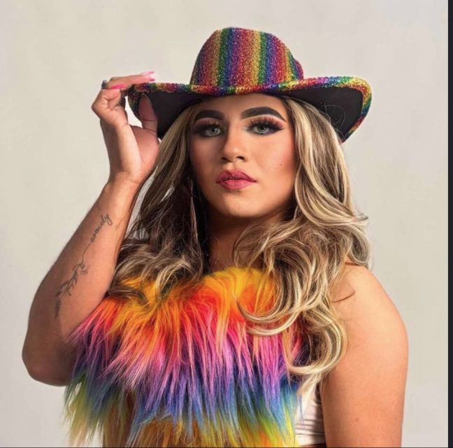 Person wearing a rainbow cowboy hat and a colorful faux fur vest, with long wavy blonde hair and makeup, posing against a plain background.