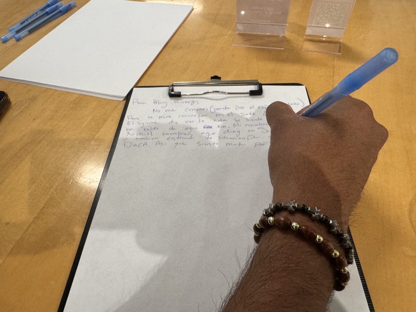 A person wearing a beaded bracelet writes on a clipboard with a blue pen; blank paper and acrylic stands are on the wooden table.