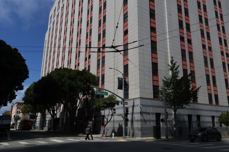 A tall, gray and pink-striped building stands at a city intersection with trees, street signs, and a few pedestrians nearby under a partly cloudy sky.