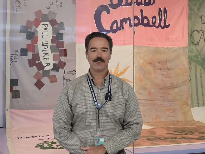 A man with a mustache wearing a checkered shirt and lanyard stands indoors in front of colorful fabric banners, one labeled "Paul Walker" and another "Campbell.