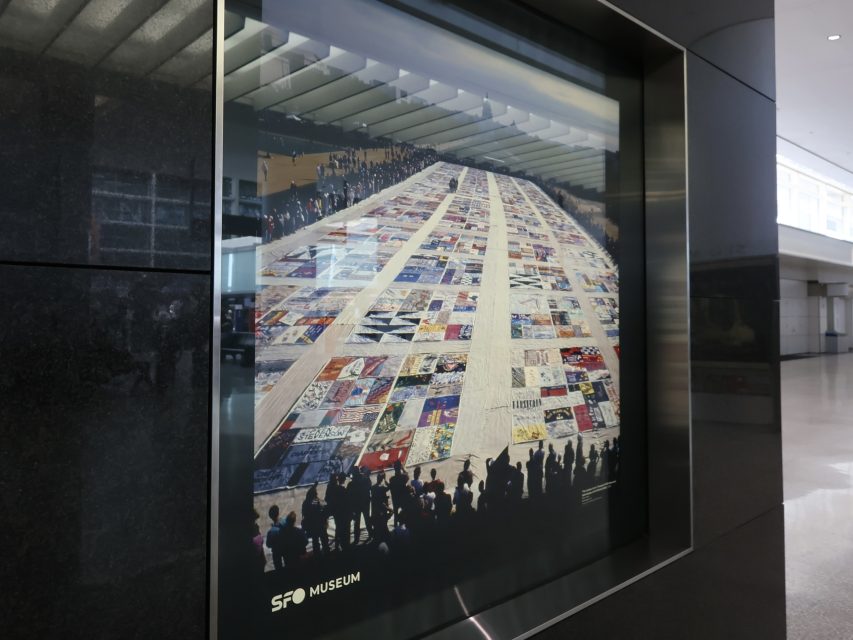 A framed photograph at SFO Museum shows a large quilt display stretching across a hall, with people gathered around its edges.