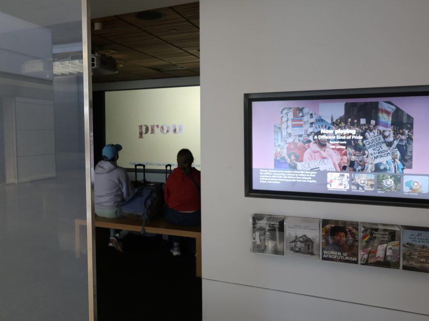 Two people sit on a bench watching a screen displaying the word “proud.” Nearby, a wall-mounted display shows images and information about protests and social movements.