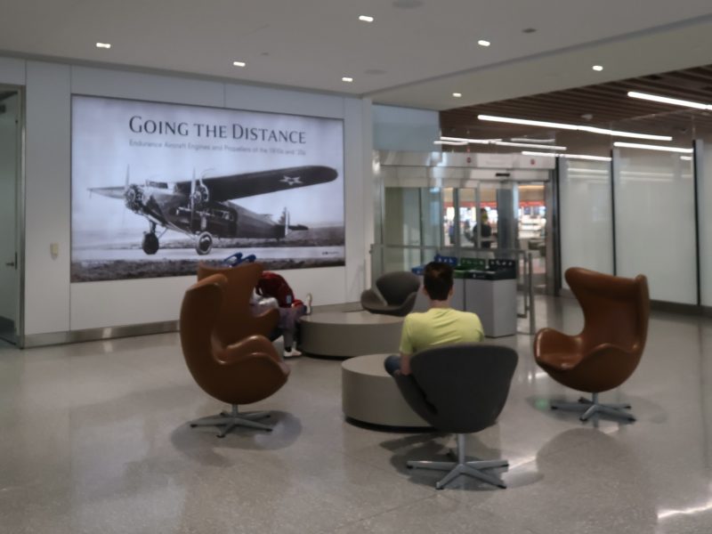 Three people sit in modern chairs near a wall mural of a vintage airplane with the text "GOING THE DISTANCE" in an airport or public space.