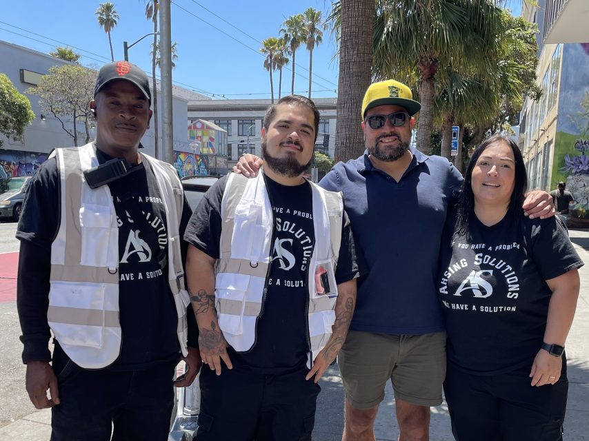 Four people stand together outdoors; three wear white vests over black "A.S. Solutions" shirts, while one wears a blue polo shirt, sunglasses, and a yellow hat. Palm trees and murals are visible.