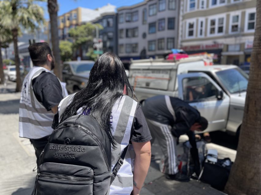 Three people in reflective vests stand and crouch near a white pickup truck parked on a city street lined with buildings. One person has a Kate Spade backpack.