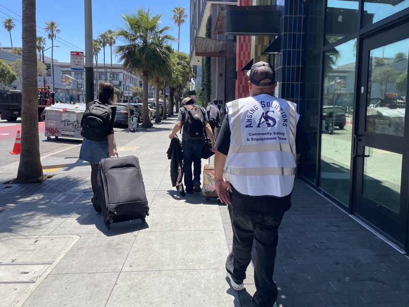 A person in a “Mission Solutions Community Safety & Engagement Team” vest walks on a sunny sidewalk behind people carrying bags and luggage.