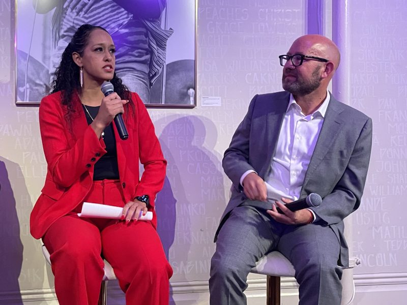 Two people sit and hold microphones during a discussion panel, with a photo and a wall covered in text in the background.
