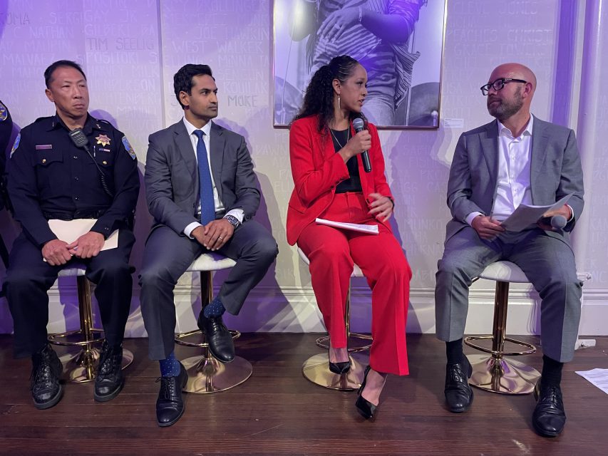 Four people sit on chairs in a panel discussion; one is in a police uniform, the others are in business attire, and one woman in red holds a microphone while speaking.