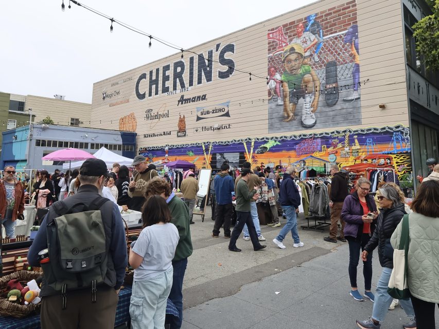 Crowd of people browsing outdoor market stalls in front of a mural-covered building with a large "Cherin's" sign.