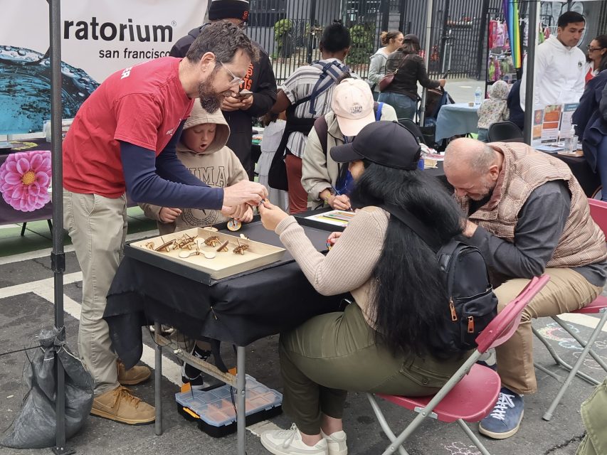 A man in a red shirt helps visitors with a hands-on science activity at an outdoor booth, while others are seated and engaged in various tasks.