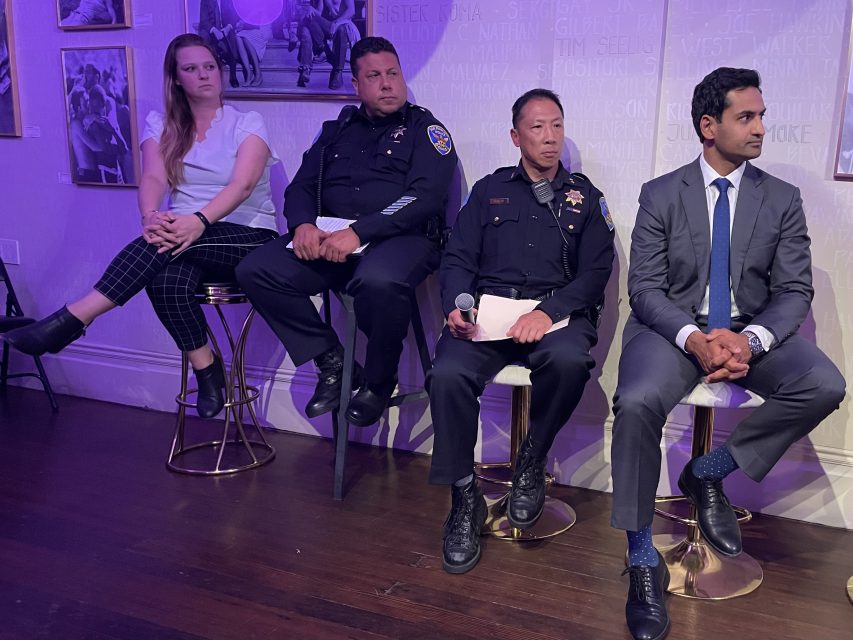Four people, including two police officers in uniform, a woman in casual attire, and a man in a suit, sit on stools against a purple-lit wall during an indoor event or panel.