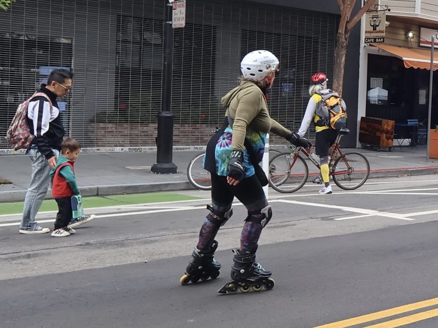 A person wearing a helmet and protective gear rollerblades on the street as a cyclist rides by and a pedestrian walks with a child on the sidewalk.