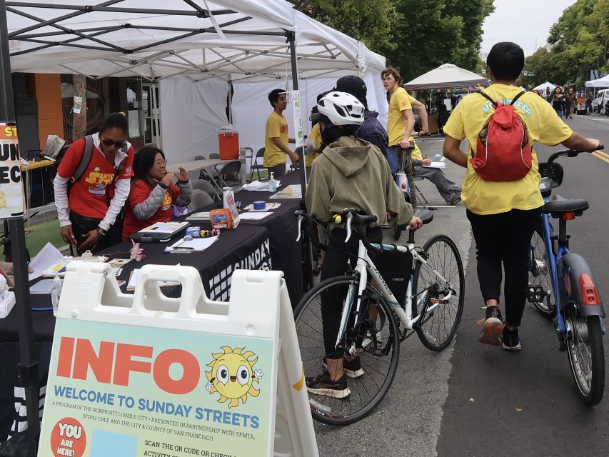 People gather around an information booth at an outdoor community event, with volunteers in yellow shirts and attendees with bicycles.