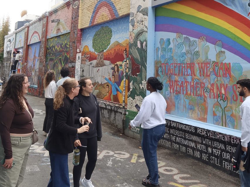 A group of people stand and look at colorful murals painted on alley walls, featuring social justice themes and the phrase "Together We Can Weather Any Storm.