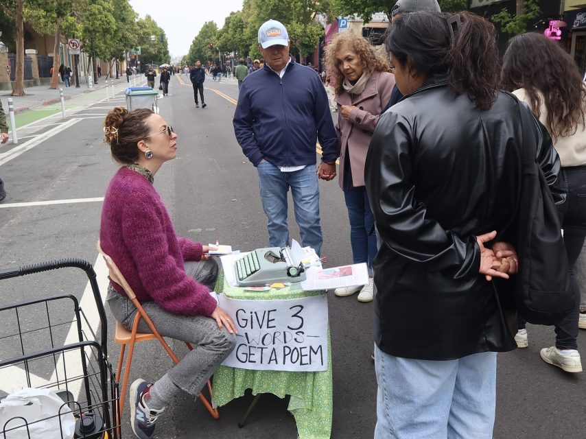A person sits at a small table with a typewriter and a sign reading “GIVE 3 WORDS GET A POEM” while talking to three people on a city street.