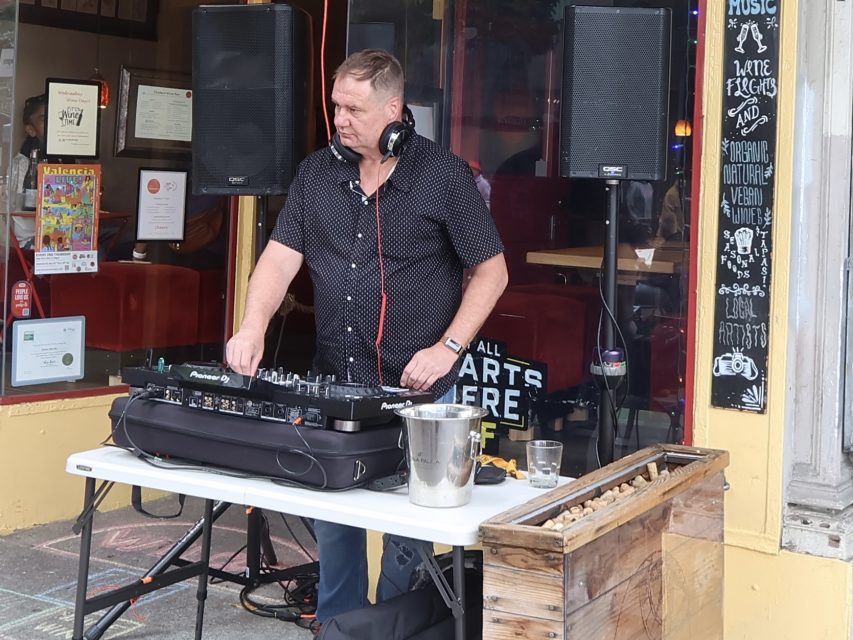 A man wearing headphones operates a DJ mixer on a sidewalk outside a cafe, with drinks and equipment on the table beside him.
