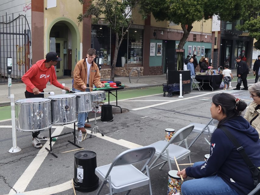 Two people play steel drums on a closed-off city street while three people seated in front watch and hold drumsticks with small practice drums.