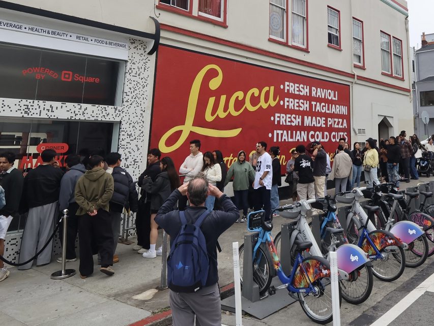 A long line of people waits outside Lucca, an Italian food shop, with bikes parked in front and a mural listing menu items on the wall.