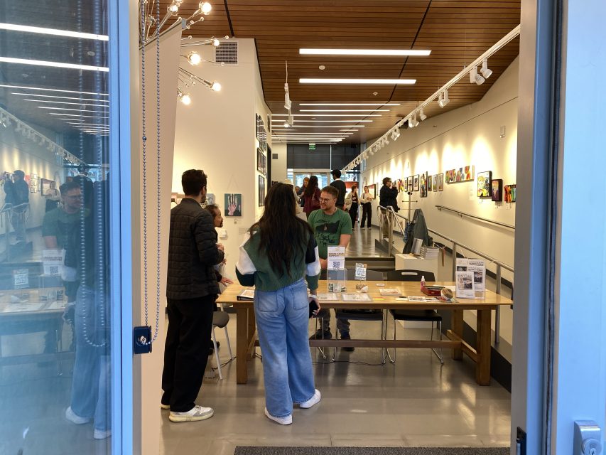 Several people stand and talk around a wooden table inside a modern art gallery with paintings displayed on white walls.