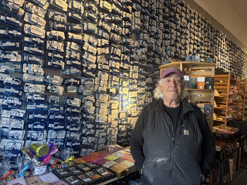 An older man stands in a shop with a wall covered in blue street sign replicas and shelves of various items by his side.