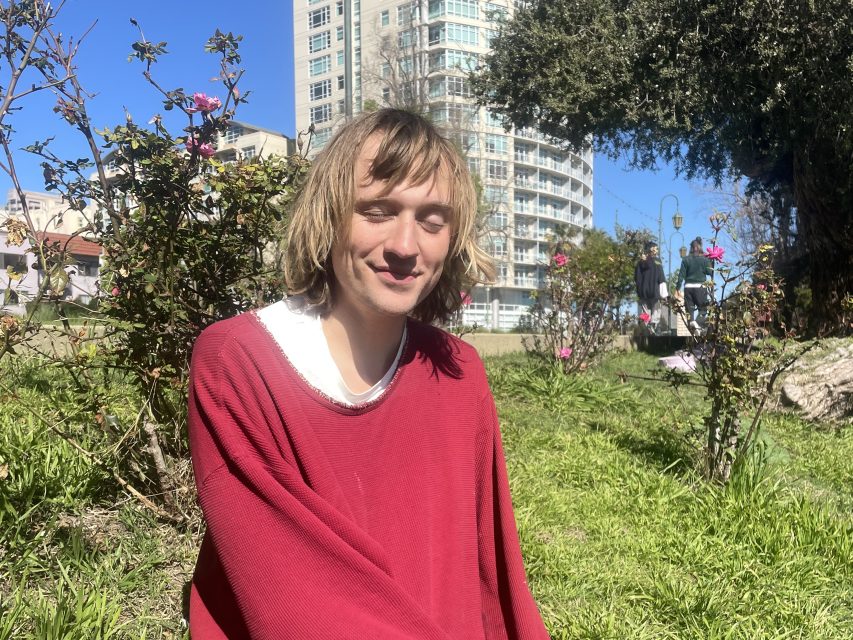 A person with shoulder-length hair wearing a red sweater sits on grass in a park, with buildings and pink flowers visible in the background on a sunny day.
