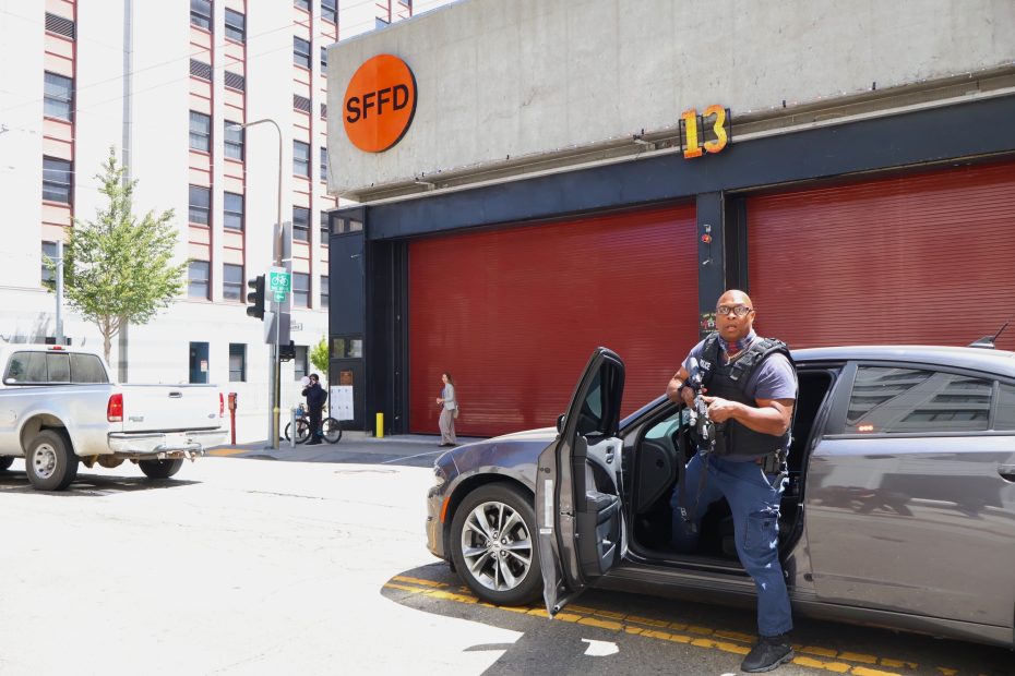 A police officer stands next to an open car door in front of a building marked “SFFD” with red garage doors and the number 13.