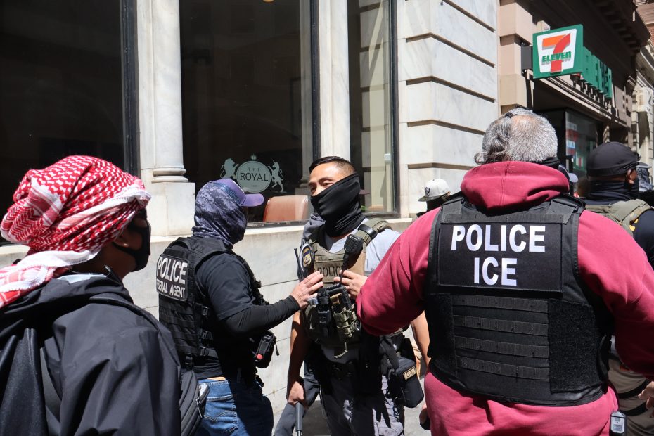 Several law enforcement officers, including ICE agents in tactical gear, stand together on a city sidewalk near a 7-Eleven store.