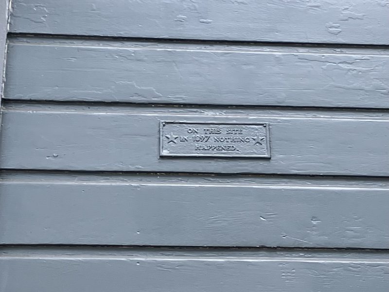 Gray wooden siding with a rectangular metal plaque mounted in the center, displaying text in raised letters. A plant is partially visible at the bottom left corner.