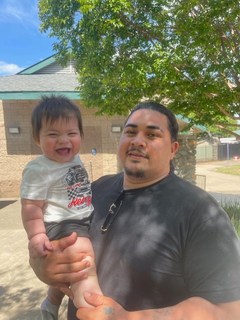 A man in a black shirt holds a smiling baby outdoors on a sunny day, with trees and a building in the background.