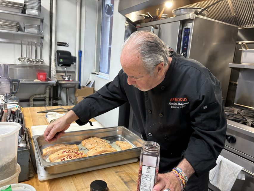 A chef in a black coat seasons salmon fillets on a tray in a commercial kitchen.