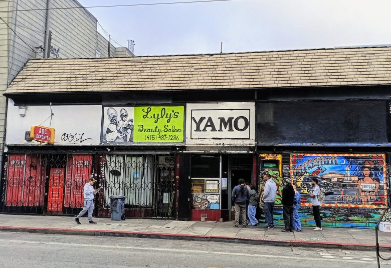 A street view of storefronts, including Lyly's Beauty Salon, Yamo restaurant, and a colorful mural, with people standing and walking on the sidewalk.
