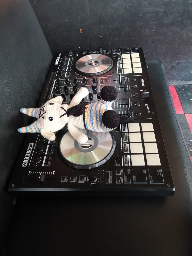 A crocheted unicorn plush toy lies on top of a DJ mixer controller, which is placed on a black leather seat.