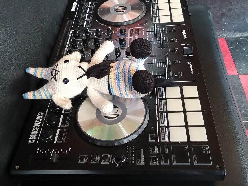 A crocheted unicorn plush toy lies on top of a DJ mixer controller, which is placed on a black leather seat.