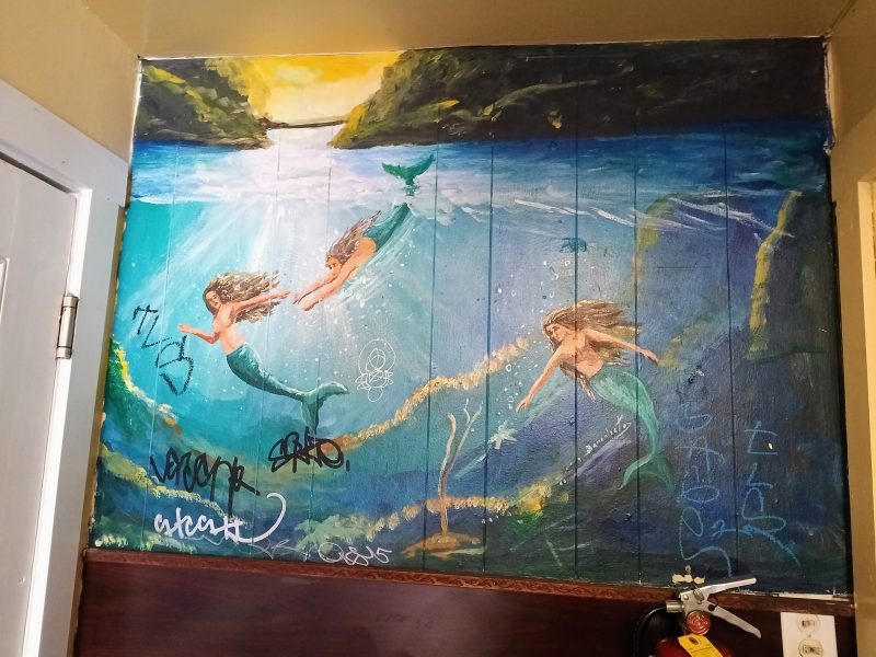 A wall mural of three mermaids swimming underwater with green tails and brown hair, partially defaced by graffiti markings and tags.