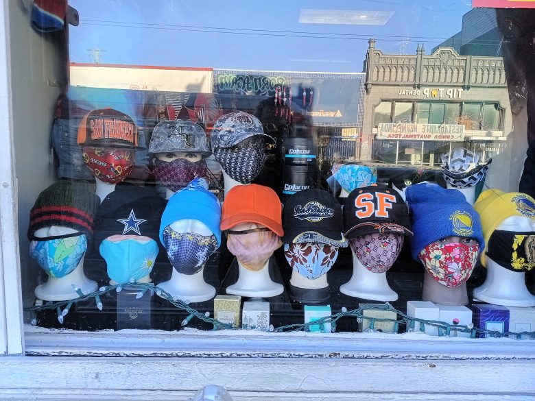 A store window display shows mannequins wearing various hats and face masks, with city buildings reflected in the glass.