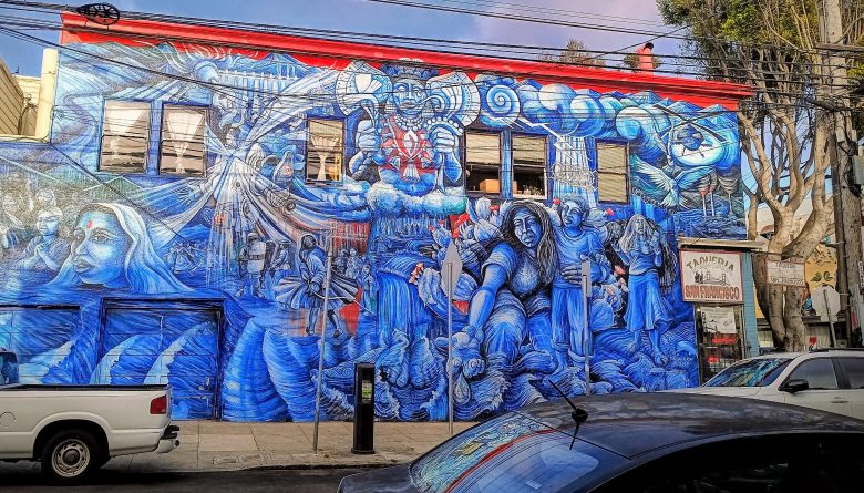 A large blue mural covers the side of a two-story building, depicting expressive human figures and intricate designs; cars are parked in front and trees line the street.