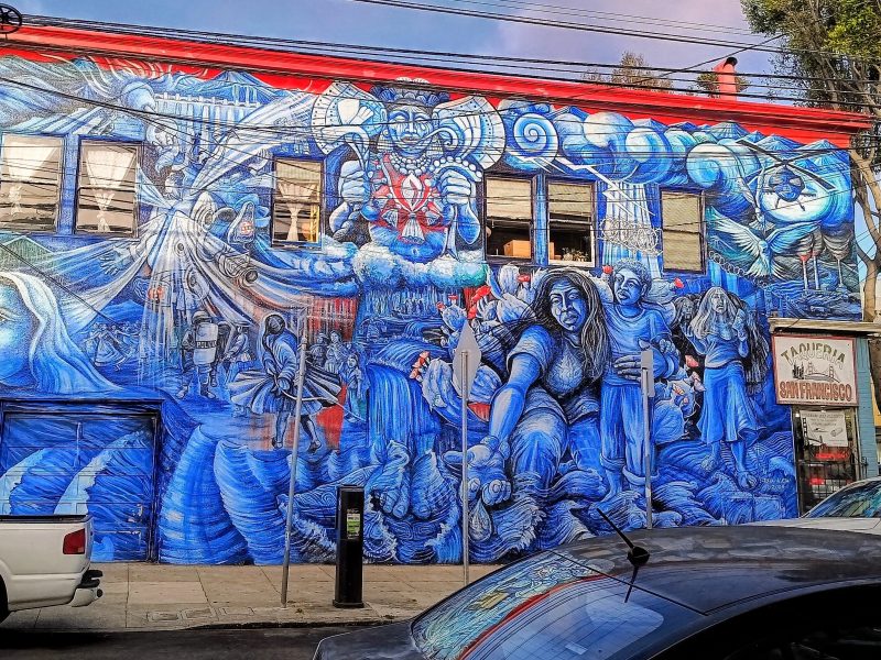 A large blue mural covers the side of a two-story building, depicting expressive human figures and intricate designs; cars are parked in front and trees line the street.