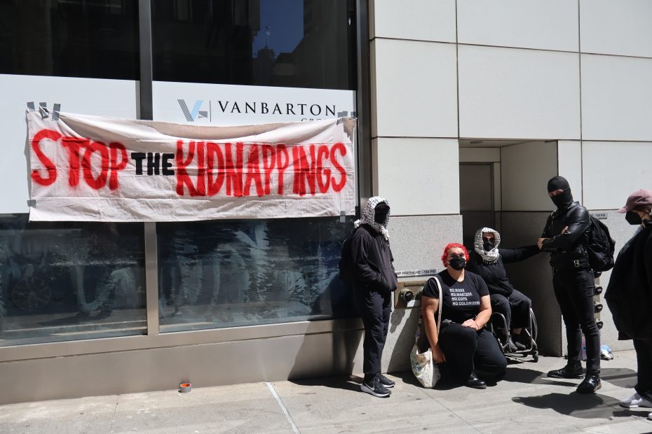 A group of people stand and sit by a building beside a large banner reading “Stop the Kidnappings” in red and black letters.