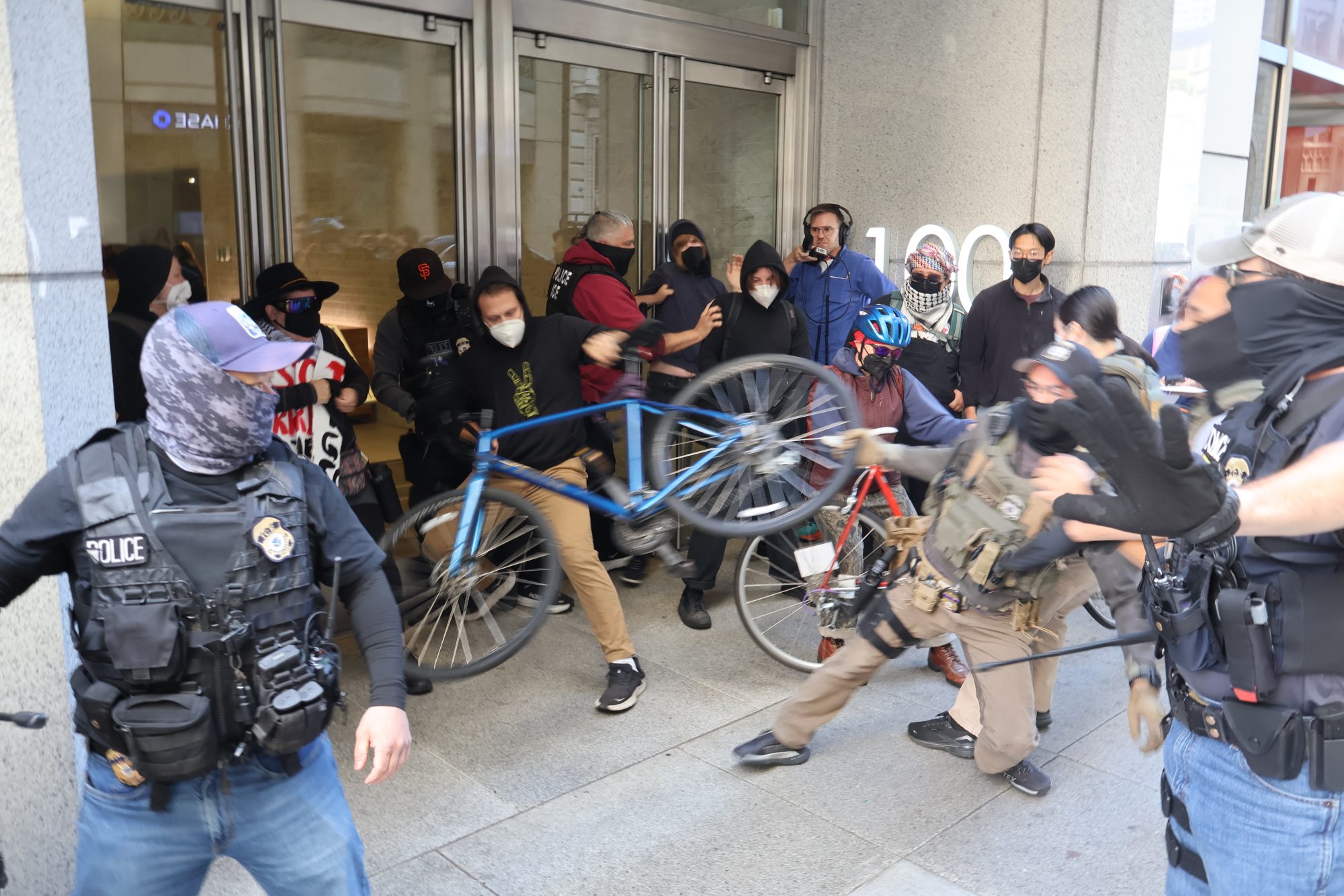 Police officers and masked individuals clash at a building entrance; one person holds up a bicycle amid the confrontation.