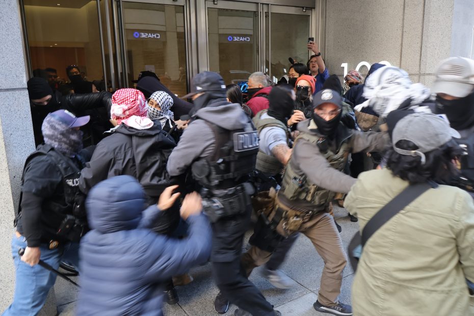 A group of masked individuals and police officers struggle in front of glass doors at a Chase bank entrance.