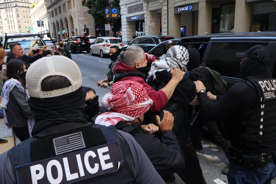 Police officers detain several people wearing keffiyehs during a street altercation in an urban area with bystanders and vehicles in the background.