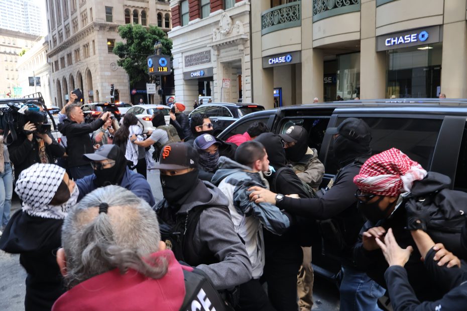 A group of masked individuals confront each other in front of a black vehicle on a city street, with bystanders observing and several people recording the scene.
