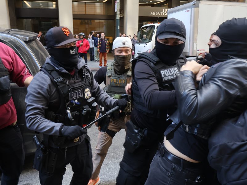Plainclothes police officers in tactical gear detain a masked individual on a city street, as bystanders watch in the background.