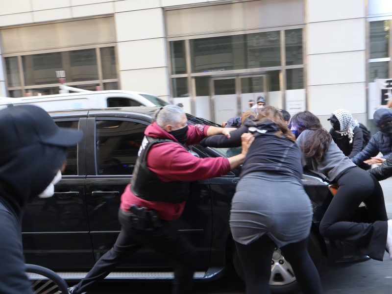 A group of people push against a black car on a city street, some wearing masks and hooded clothing, next to a building with large windows.