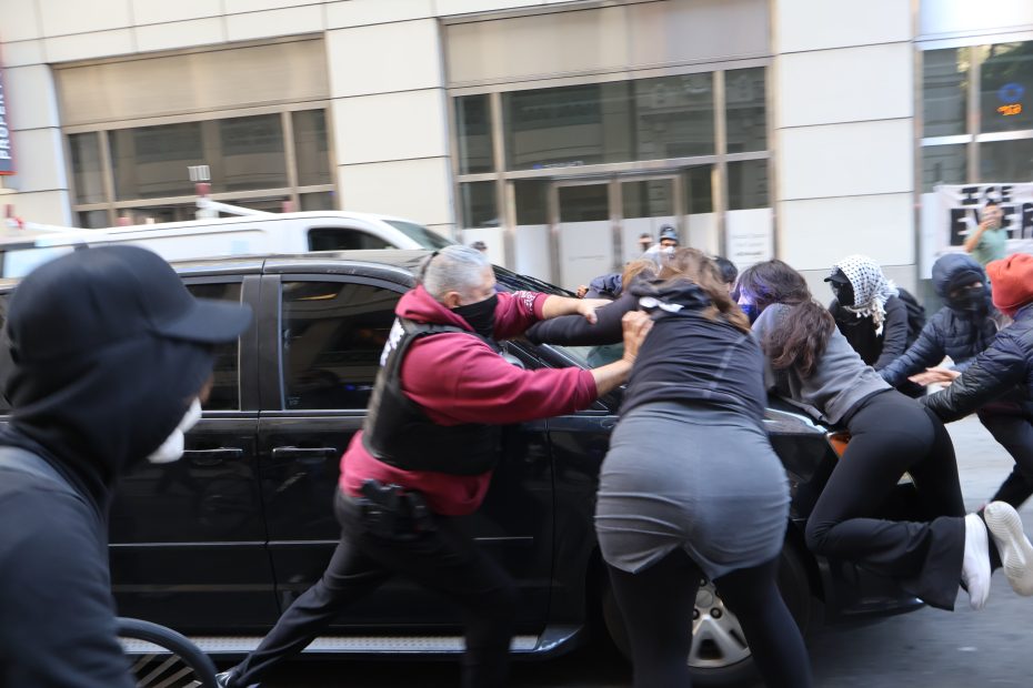 A group of people push against a black car on a city street, some wearing masks and hooded clothing, next to a building with large windows.