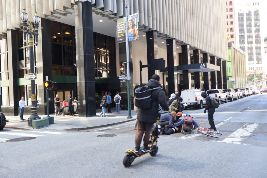 People attend to a person on the ground next to a fallen bicycle at a city street intersection while a man rides by on a scooter. Pedestrians and parked vehicles are visible nearby.