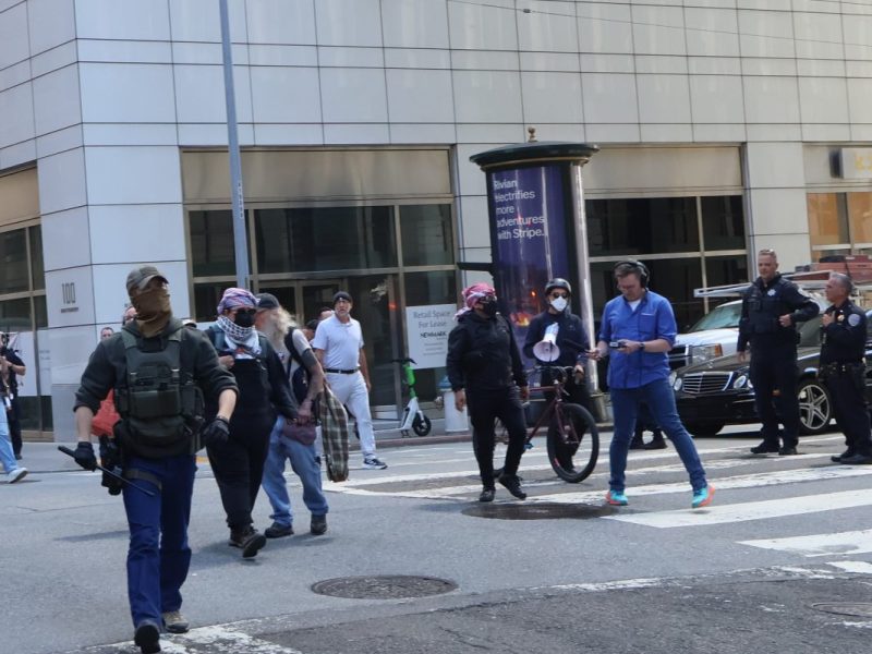A group of people, some wearing tactical gear and masks, walk across a crosswalk in an urban area with tall buildings and parked cars.