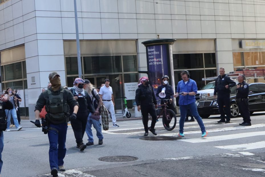 A group of people, some wearing tactical gear and masks, walk across a crosswalk in an urban area with tall buildings and parked cars.